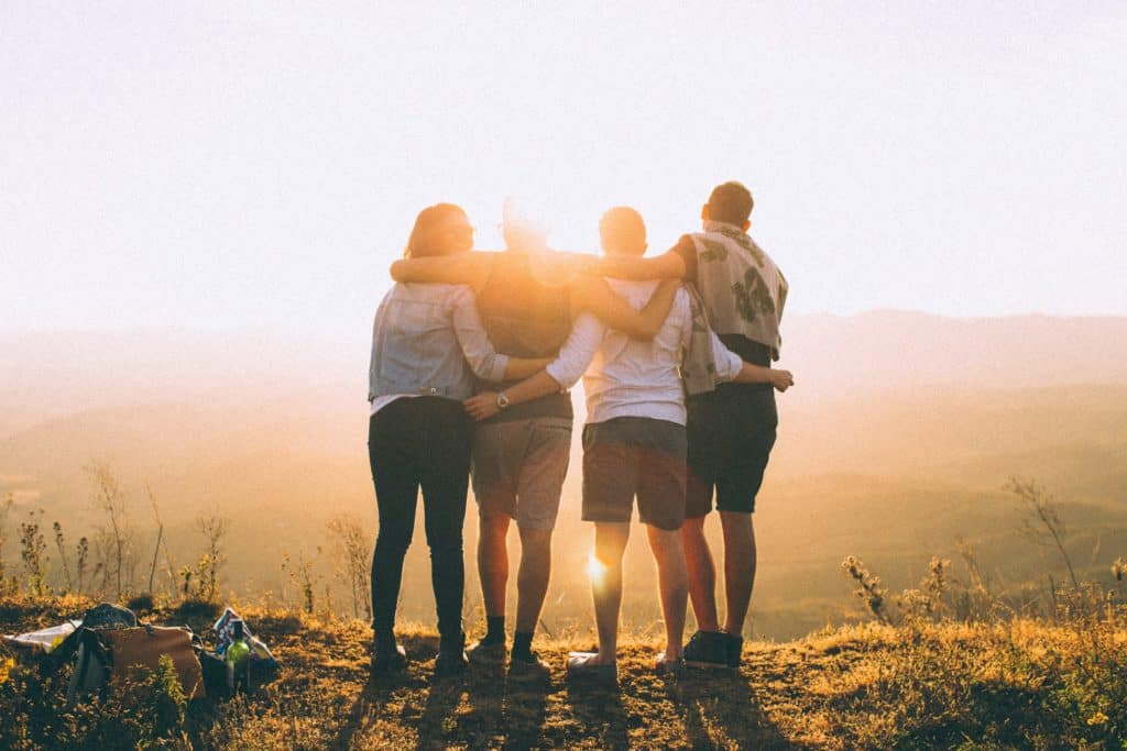 Young people stand looking off of a ledge into a sunset with their arms wrapped around each other as part of an alumni event.