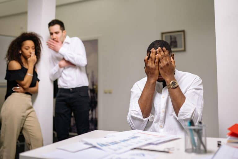 frustrated man at work covering his face while other employees talk.